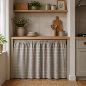 Könnte beinhalten: Kücheninterieur mit einem grau-weiß karierten Vorhang. Holzregale beherbergen Pflanzen, Gläser und einen gerahmten Druck. Eine Vase mit Blumen und Tellern steht auf der Theke. Ein Holzschneidebrett und Utensilien sind sichtbar. Ein gewebter Teppich liegt auf dem Boden.