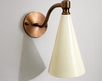 May include: A wall-mounted sconce with a conical off-white shade. The fixture features a copper-colored circular base and a curved arm in a bronze finish. The sconce is designed for indoor lighting.