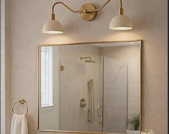 May include: A bathroom vanity set featuring a rectangular mirror with a gold frame, a two-light wall sconce with curved gold arms and cream dome shades, a gold faucet, a white towel on a gold ring, and a small vase with white flowers.