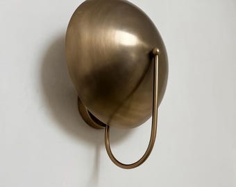 May include: A bronze-colored wall sconce with a rounded, egg-like shade and a curved handle. The sconce is mounted on a white wall, creating a contrast with its warm metallic finish. The design is modern and minimalist.