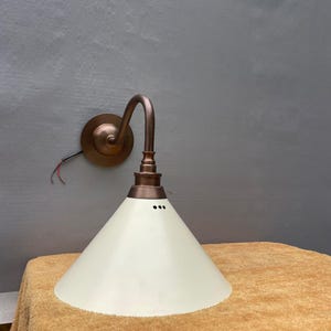 May include: A copper-finished wall light with an off-white, conical shade. The fixture has a curved arm and a circular base. The shade has three small holes near the top. The light is designed for wall mounting.