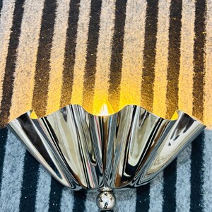 Op de afbeelding: Een wandlamp met een gepolijste, chromen, gegolfde kap in de vorm van een kom die het licht reflecteert. Het armatuur bevat een centraal bolvormig accent en een zwart netsnoer, met een warm gloeiende lamp zichtbaar in de kap.