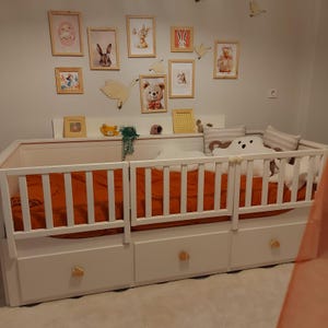 May include: A white toddler bed with an orange duvet and white safety rails. The bed has three drawers with gold-coloured knobs. Above the bed, there are framed pictures of animals and decorative elements on the wall.