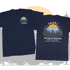May include: A navy blue t-shirt with a graphic of a mountain range and a yellow sun. The text "Kaleb's Bachelor Weekend 2023" is printed on the shirt. The text "Bachelor Weekend North Carolina" and "Good friends, good beer, good times" is also printed on the shirt.