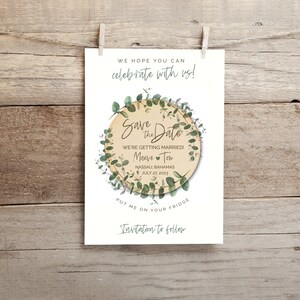 May include: Save the Date card with a green eucalyptus wreath around a wooden circle with the text "Save the Date" and the names "Maeve" and "Tom" with a heart between them. The text also includes "We're getting married!" and "Nassau, Bahamas July 27, 2023". The card also has the text "Put me on your fridge" and "Invitation to follow".