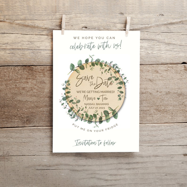 May include: Save the Date card with a green eucalyptus wreath around a wooden circle with the text "Save the Date" and the names "Maeve" and "Tom" with a heart between them. The text also includes "We're getting married!" and "Nassau, Bahamas July 27, 2023". The card also has the text "Put me on your fridge" and "Invitation to follow".