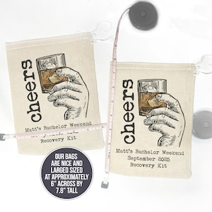 May include: Two beige canvas bags with drawstring closures. Each bag features a black and white illustration of a hand holding a glass of whiskey with ice. The text "cheers" is printed vertically on the bag. The text "Matt's Bachelor Weekend" and "Recovery Kit" is printed on the bottom of each bag. The bags are approximately 6 inches wide and 7.8 inches tall.