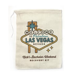May include: A white canvas bag with a drawstring closure. The bag features a retro-style illustration of the Las Vegas sign with the text "Welcome to Fabulous Las Vegas, Nevada". Below the sign, the text reads "Nick's Bachelor Weekend Recovery Kit".
