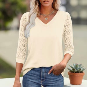 May include: Cream-coloured V-neck top with three-quarter sleeves featuring a delicate lace pattern. The top is paired with blue jeans. A small potted succulent is visible in the bottom right corner.