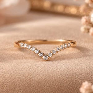 Peut inclure: Une bague en forme de V en or rose, ornée d'une série de petites pierres rondes et transparentes serties le long de l'anneau en chevron. La bague présente une finition métallique polie et un design symétrique se terminant par une pointe.