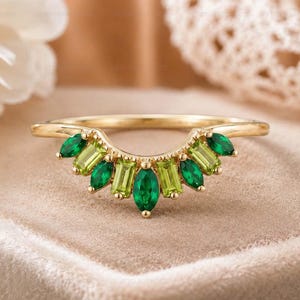 Peut inclure: Une bague en or incurvée ornée d'une rangée de pierres précieuses alternant taille marquise et taille baguette dans des tons vert émeraude et vert lime. Les pierres sont disposées symétriquement le long de l'arc de la fine bague en or poli.