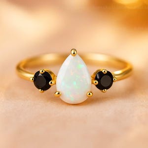 May include: A gold ring featuring a pear-shaped white opal center stone, flanked by two round black gemstones. The ring has a delicate band and small gold accents around the stones. A beautiful piece of jewelry.