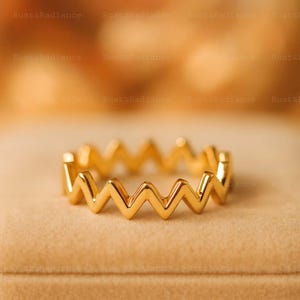 Puede incluir: Un anillo dorado con diseño en zigzag. El anillo presenta un patrón en forma de V repetido, creando un efecto ondulado continuo. Se muestra sobre una superficie beige, resaltando su brillo metálico.