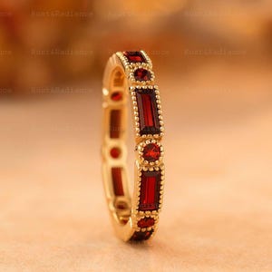 May include: A gold ring featuring alternating rectangular and round red gemstones. The ring has a detailed design with small, raised dots around the gemstones. The ring is set against a blurred, warm-toned background.