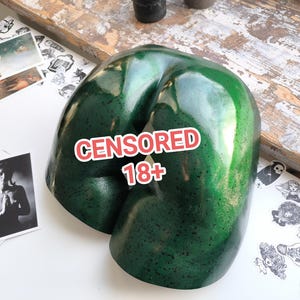 Könnte beinhalten: Eine glänzende, dunkelgrüne Skulptur mit einer geschwungenen, abstrakten Form. Die Skulptur hat eine strukturierte Oberfläche und ist mit dem Text "CENSORED 18+" in Rot und Weiß beschriftet. Das Objekt befindet sich auf einer weißen Oberfläche mit Kunstdrucken und Aufklebern.