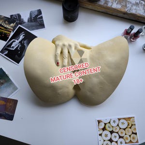 May include: A beige sculpture with a hand resting on it. The hand has a red painted fingernail. Several photographs and bottles of nail varnish are scattered around the sculpture.
