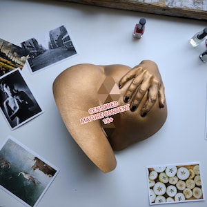 May include: A gold-coloured sculpture of a human form with a hand covering the form. The hand has black nail polish. Several small photographs and bottles of nail polish are scattered around the sculpture.