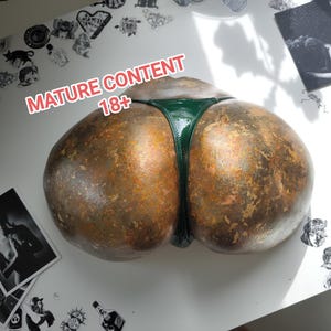 May include: A sculpture of a derriere with a green thong. The sculpture has a textured, bronze-like appearance. The text "MATURE CONTENT 18+" is visible in the image.