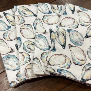 May include: Set of six white cotton napkins with a watercolor design of blue and green oysters. The napkins are folded and stacked on a wooden surface.