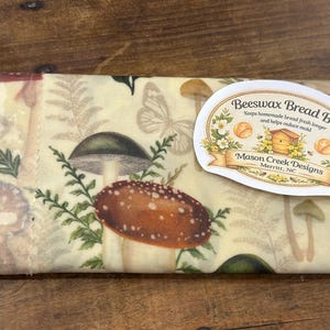 May include: Beeswax bread bag with a cream background and a mushroom and fern pattern. The bag has a label that reads "Beeswax Bread Bags" and "Mason Creek Designs, Merritt, NC."