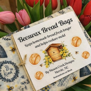 May include: Beeswax bread bags with a floral and bee design. The bags are wrapped with a twine tie and surrounded by red, pink, and yellow tulips. The label reads "Beeswax Bread Bags" and "Keeps homemade bread fresh longer and helps reduce mold."