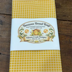 May include: A yellow and white gingham beeswax bread bag wrapped in a white paper band. The band features the text Beeswax Bread Bags, Keeps homemade bread fresh longer and helps reduce mold, Mason Creek Designs, and Merritt, NC.