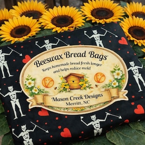 May include: A black beeswax bread bag with a repeating skeleton and heart pattern. The bag has a central oval label with the text "Beeswax Bread Bags" and "Mason Creek Designs, Merritt, NC". Sunflowers and greenery surround the bag.