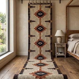 May include: A long, rectangular woven rug featuring a repeating geometric pattern in shades of rust orange, black, and cream. The rug is suspended from a horizontal wooden rod by metal clips, extending from the wall down onto the wooden floor.