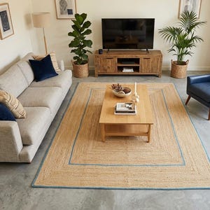 May include: A living room scene featuring a large, rectangular jute rug with a light blue border. The room includes a light gray sofa with navy blue and patterned throw pillows, a wooden coffee table, and a blue armchair. Decorative plants and artwork complete the space.