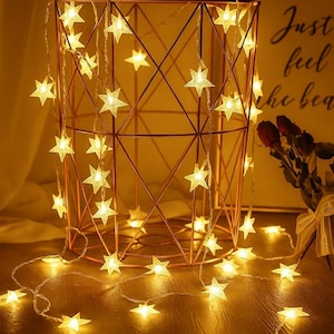 May include: A decorative copper wire basket with a geometric design is draped with a string of glowing star-shaped lights. The lights are warm white and create a cozy ambiance. The background includes a sign with the words "Just feel the bea".