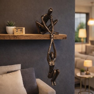 May include: A bronze sculpture of two figures, one seated on a wooden shelf and the other hanging below, holding onto the first figure's hands. The sculpture is a decorative piece with a modern aesthetic. A small plant and a calendar are also on the shelf.