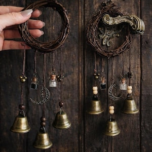 May include: Two rustic dream catchers with a circular woven twig frame. Each features hanging bronze bells, glass vials, crescent moon charms, and wooden beads. The background is a dark wooden surface.