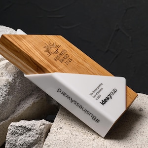 May include: A wooden business award with a white triangular base. The award features the text "BUSINESS AWARD 2023" and the hashtag "#BusinessAward". The award is set against a textured background.