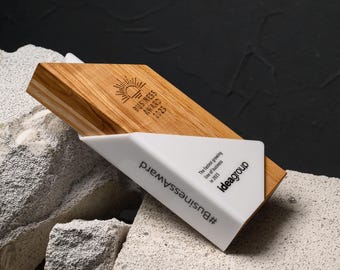 May include: A wooden business award with a white triangular base. The award features the text "BUSINESS AWARD 2023" and the hashtag "#BusinessAward". The award is set against a textured background.