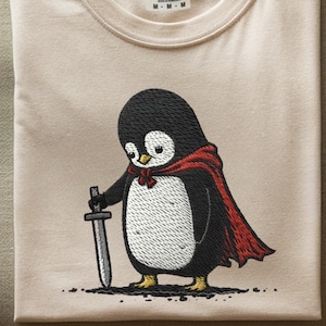 May include: A folded, beige t-shirt featuring an embroidered cartoon penguin wearing a red cape and holding a sword. The penguin has a black head, white chest, and yellow feet. The shirt has a Comfort Colors tag.