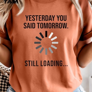 May include: Burnt orange t-shirt with the text "YESTERDAY YOU SAID TOMORROW. STILL LOADING..." and a loading symbol. The shirt has rolled-up sleeves and a crew neck.
