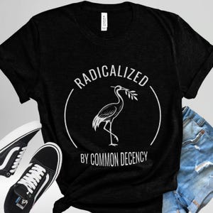 May include: Black t-shirt with a white graphic of a heron holding a sprig, encircled by the words "RADICALIZED" and "BY COMMON DECENCY". The shirt is paired with black sneakers and blue denim shorts.