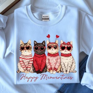 May include: A light blue sweatshirt featuring four cartoon cats wearing heart-shaped sunglasses and sweaters. The cats have heart-shaped accents and the words "Happy Meowentines" are printed in red script below the cats.