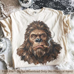 May include: A cream-colored short-sleeve t-shirt featuring a detailed, painterly graphic of a brown, furry creature resembling a sasquatch or ape. The creature is depicted with a cigar in its mouth, with a thin trail of smoke rising upward.