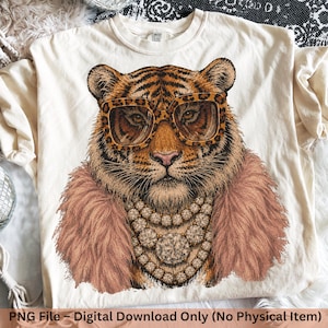 Könnte beinhalten: Ein cremefarbenes T-Shirt mit einer grafischen Illustration eines Tigers, der eine Brille mit Leopardenmuster, einen rosa Kunstfellkragen und eine mehrlagige Strasshalskette trägt. Das T-Shirt hat einen Rundhalsausschnitt und kurze Ärmel.