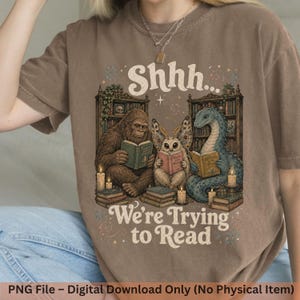 Può includere: Una t-shirt marrone a maniche corte con una stampa grafica di Bigfoot, una creatura simile a una falena e un serpente marino che leggono libri in una biblioteca. Il testo recita Shhh... We're Trying to Read. Il design include librerie e candele.