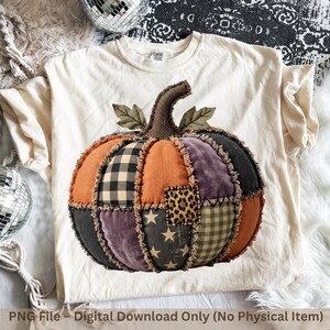 Peut inclure: Un t-shirt couleur crème arborant un grand motif central de citrouille en patchwork. La citrouille est composée de diverses textures et motifs de tissus, notamment orange, noir, violet, imprimé léopard, carreaux vichy et étoiles.