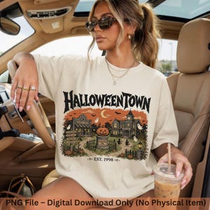 May include: A cream-colored crewneck t-shirt featuring a graphic print of a spooky town with the text HALLOWEENTOWN and EST. 1998. The illustration includes a pumpkin on a pedestal, ghosts, buildings, and trees in shades of orange, black, and brown.