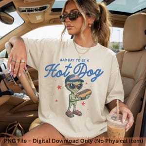 May include: A cream-colored t-shirt featuring a graphic of a green alien wearing a US flag baseball cap, red sunglasses, and red sandals. The alien holds a hot dog. The text reads "BAD DAY TO BE A HOT DOG" in blue script with red stars.