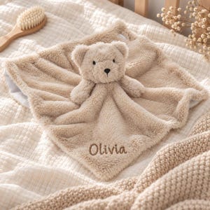 May include: A beige plush security blanket featuring a centred teddy bear head with embroidered facial features. The soft, fuzzy fabric has the name Olivia embroidered in brown thread on one corner, creating a tactile and comforting item.
