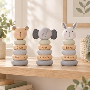 Puede incluir: Tres juguetes apilables de madera con forma de oso, elefante y conejo. Cada juguete tiene anillos apilados en tonos grises y madera natural. Los anillos centrales están grabados con los nombres Olivia, Noah y Emma, y las fechas 10.03.2026, 18.06.2026 y 24.08.2026.