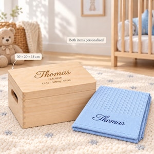 Op de afbeelding: Een lichtgekleurde houten aandenkenbox van 30 x 20 x 14 cm, gegraveerd met de naam Thomas, een datum, tijd, gewicht en lengte. Daarnaast ligt een gevouwen lichtblauwe gebreide deken geborduurd met de naam Thomas in donkerblauw cursief schrift.