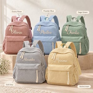 Pode incluir: Cinco mochilas de veludo cotelê nas cores rosa antigo, azul pó, verde sálvia, cinza suave e creme. Cada mochila possui um bolso frontal com zíper, bolsos laterais, puxadores de corda branca e os nomes bordados Olivia, Matteo, Charlie, Alexander e Maxime.