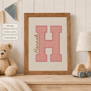 Op de afbeelding: Een ingelijst borduurwerk met een grote roze letter H met een ruitpatroon en de naam Hannah gestikt in bruin cursief garen op een lichtbeige stoffen achtergrond, gepresenteerd in een houten lijst met de tekst A4-formaat.