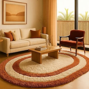 Puede incluir: Una alfombra ovalada de pelo largo con un centro crema y dos bordes concéntricos en tonos naranja. La alfombra está en un salón con un sofá, un sillón y una mesa de centro. El ambiente es cálido y acogedor.